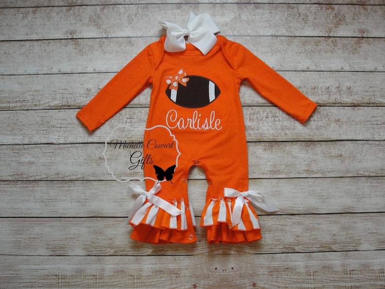May include: Orange and white baby onesie with a brown football and white bow on the front. The onesie has a white ruffle trim and the name "Carlisle" is embroidered on the front.