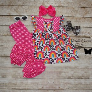 Puede incluir: Un conjunto de leggings de volantes de lunares rosa y blanco con un vestido de estampado de arco&iacute;ris y leopardo y una diadema de lazo rosa. El conjunto es para una ni&ntilde;a peque&ntilde;a.