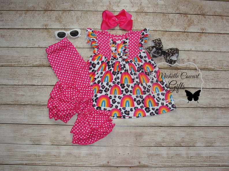 Puede incluir: Un conjunto de leggings de volantes de lunares rosa y blanco con un vestido de estampado de arco&iacute;ris y leopardo y una diadema de lazo rosa. El conjunto es para una ni&ntilde;a peque&ntilde;a.