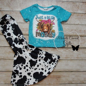 Pode incluir: Conjunto de camiseta e cal&ccedil;as boca de sino. A camiseta turquesa apresenta um gr&aacute;fico de uma vaca das Terras Altas com o texto "Just a little moody". As cal&ccedil;as t&ecirc;m uma estampa de vaca em preto e branco. Conjunto para crian&ccedil;as.