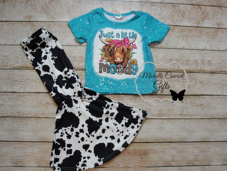 Pode incluir: Conjunto de camiseta e cal&ccedil;as boca de sino. A camiseta turquesa apresenta um gr&aacute;fico de uma vaca das Terras Altas com o texto "Just a little moody". As cal&ccedil;as t&ecirc;m uma estampa de vaca em preto e branco. Conjunto para crian&ccedil;as.