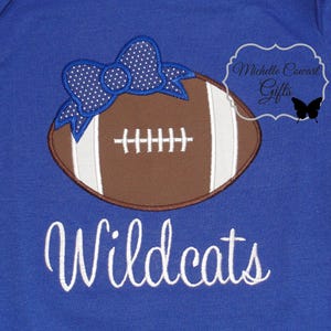 May include: A royal blue t-shirt with a brown and white football with a blue and white polka dot bow on top. The text "Wildcats" is embroidered in white below the football.