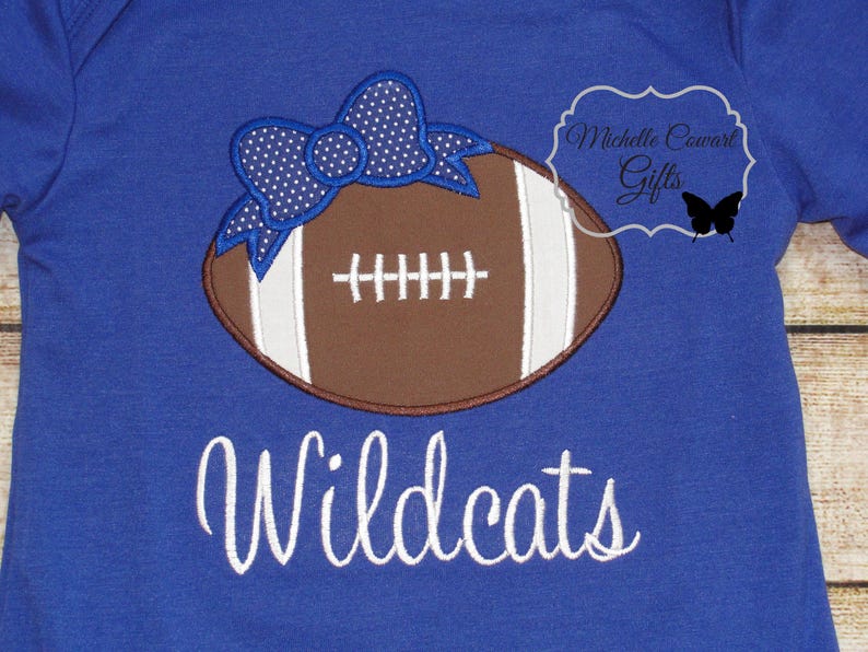 May include: A royal blue t-shirt with a brown and white football with a blue and white polka dot bow on top. The text "Wildcats" is embroidered in white below the football.