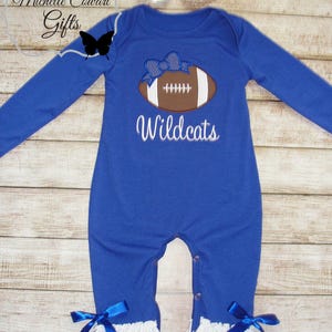 May include: Blue long-sleeve baby onesie with a white ruffle trim and blue bows. The onesie features an embroidered brown football with a blue bow on top and the text "Wildcats".