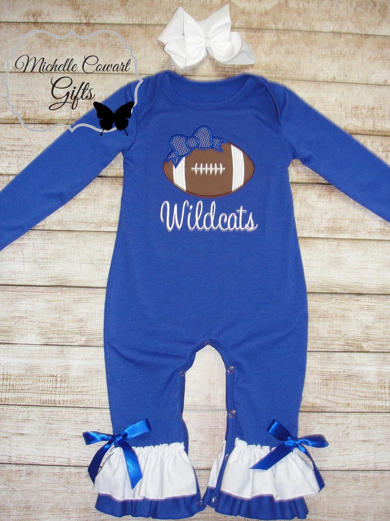 May include: Blue long-sleeve baby onesie with a white ruffle trim and blue bows. The onesie features an embroidered brown football with a blue bow on top and the text "Wildcats".
