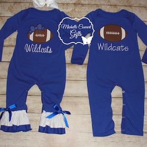 May include: Two blue baby onesies with white ruffles at the bottom. One onesie has a football and the text "Wildcats" embroidered on it. The other onesie has a football with a bow on it and the text "Wildcats" embroidered on it.