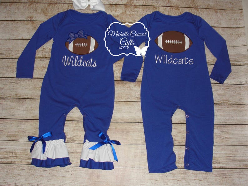 May include: Two blue baby onesies with white ruffles at the bottom. One onesie has a football and the text "Wildcats" embroidered on it. The other onesie has a football with a bow on it and the text "Wildcats" embroidered on it.