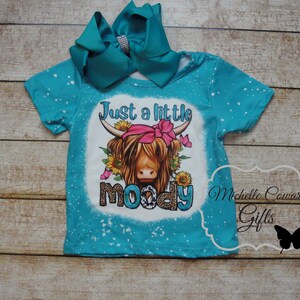 Pode incluir: Camiseta turquesa com um gr&aacute;fico de uma vaca Highland usando uma bandana rosa e cercada por girass&oacute;is e borboletas. O texto diz "Just a little moody". Um grande la&ccedil;o turquesa com um centro de strass est&aacute; preso.