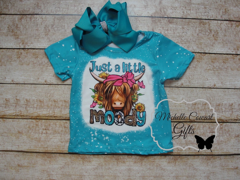 Pode incluir: Camiseta turquesa com um gr&aacute;fico de uma vaca Highland usando uma bandana rosa e cercada por girass&oacute;is e borboletas. O texto diz "Just a little moody". Um grande la&ccedil;o turquesa com um centro de strass est&aacute; preso.