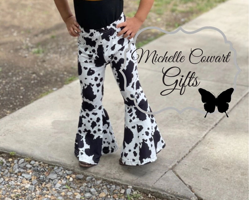 Pode incluir: Cal&ccedil;as de boca de sino com estampa de vaca em preto e branco. As cal&ccedil;as t&ecirc;m um design de perna flare e um padr&atilde;o repetido de manchas pretas sobre um fundo branco. O texto "Michelle Cowart Gifts" &eacute; vis&iacute;vel.