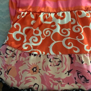 May include: A tiered dress featuring a striped pink and orange bodice. The layers below showcase orange fabric with white swirls, a floral print in pink and peach tones, and a black and white patterned hemline. The dress has a ruffled design.