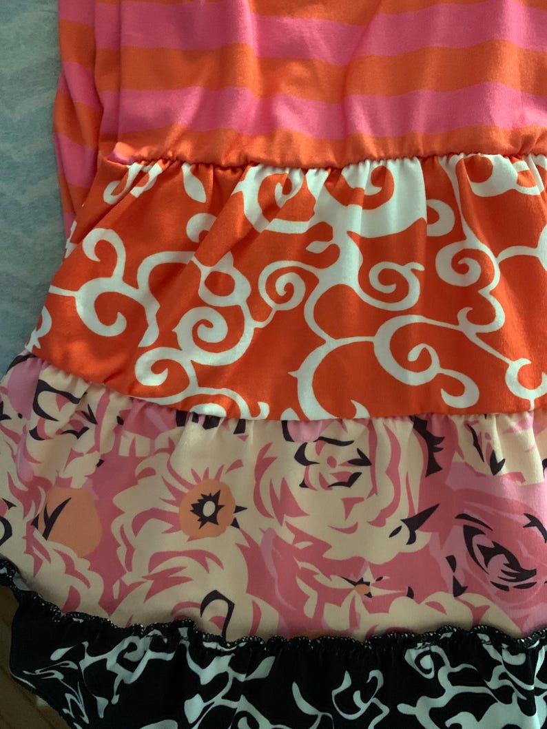May include: A tiered dress featuring a striped pink and orange bodice. The layers below showcase orange fabric with white swirls, a floral print in pink and peach tones, and a black and white patterned hemline. The dress has a ruffled design.