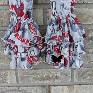 May include: Two ruffled leg warmers with a repeating pattern of red, white, and gray text and graphics, including the word "ALABAMA". The leg warmers have multiple layers of ruffles.