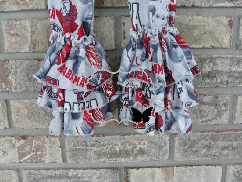 May include: Two ruffled leg warmers with a repeating pattern of red, white, and gray text and graphics, including the word "ALABAMA". The leg warmers have multiple layers of ruffles.