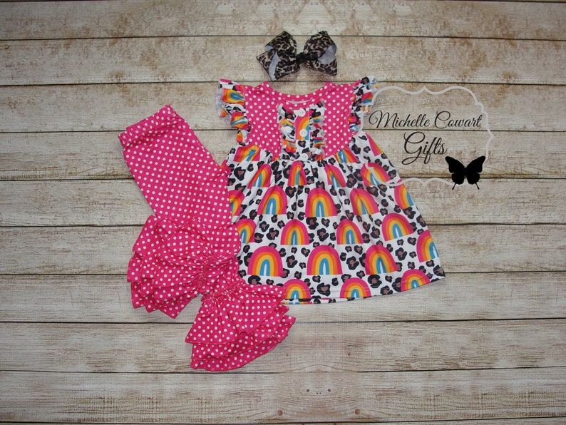 Puede incluir: Un conjunto de leggings con volantes de lunares rosa y blanco con un vestido de estampado de arco&iacute;ris y leopardo. El vestido tiene cierre de bot&oacute;n y mangas con volantes. El conjunto se combina con un lazo marr&oacute;n y negro.