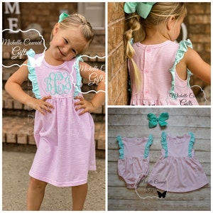 May include: A pink and white striped dress with a mint green ruffle trim and a monogram on the front. The dress has a button closure in the back. The dress is shown on a young girl. The image also shows a matching mint green headband and a pink and white striped bodysuit with a mint green ruffle trim.