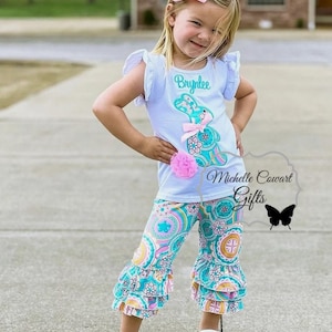 May include: A little girl wearing a white shirt with a turquoise bunny applique and the name "Brylee" embroidered on it. She is also wearing turquoise and pink patterned pants with ruffles. The pants have a paisley design.