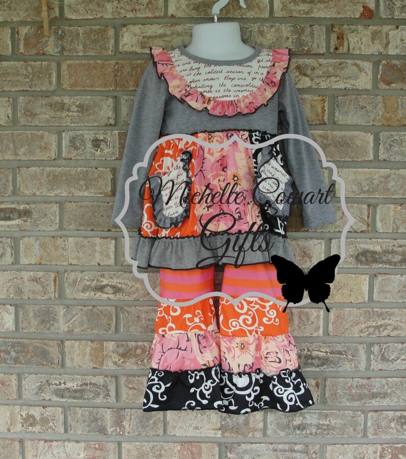 May include: A gray long-sleeved top and tiered pants set for girls. The top has a ruffled collar with floral and script details. The pants feature orange, pink, and black patterned tiers. The set is displayed on a mannequin.
