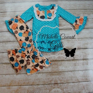 Puede incluir: Un conjunto turquesa con lunares blancos con tem&aacute;tica de calabazas. La parte superior de manga larga tiene un dobladillo con volantes y un cuello de encaje blanco. Los pantalones a juego presentan un estampado de calabaza en naranja, negro y azul. El texto "Michelle Cowart Gifts" es visible.