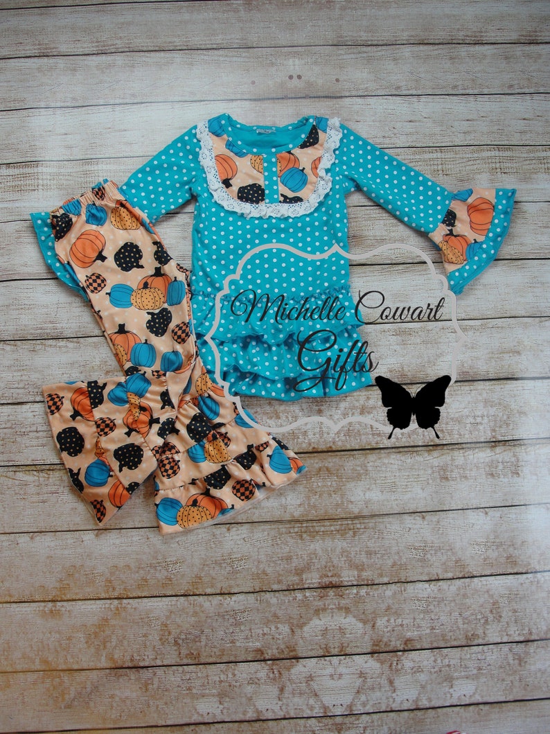 Puede incluir: Un conjunto turquesa con lunares blancos con tem&aacute;tica de calabazas. La parte superior de manga larga tiene un dobladillo con volantes y un cuello de encaje blanco. Los pantalones a juego presentan un estampado de calabaza en naranja, negro y azul. El texto "Michelle Cowart Gifts" es visible.