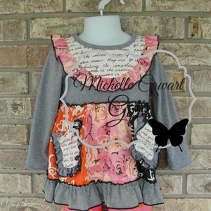 May include: A gray long-sleeved top with a floral and text-printed design. The top has a ruffled collar and hem, with orange and black accents. The text "Michelle Cowart" is visible.