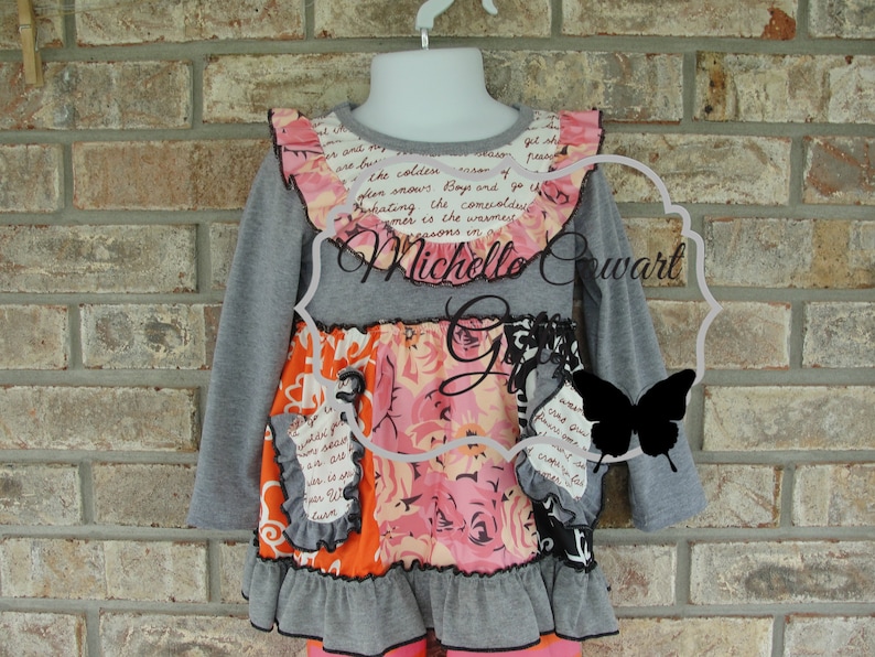 May include: A gray long-sleeved top with a floral and text-printed design. The top has a ruffled collar and hem, with orange and black accents. The text "Michelle Cowart" is visible.