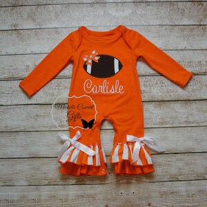 May include: Orange and white baby onesie with a football and bow design. The onesie has a ruffled bottom and says "Carlisle" on the front.
