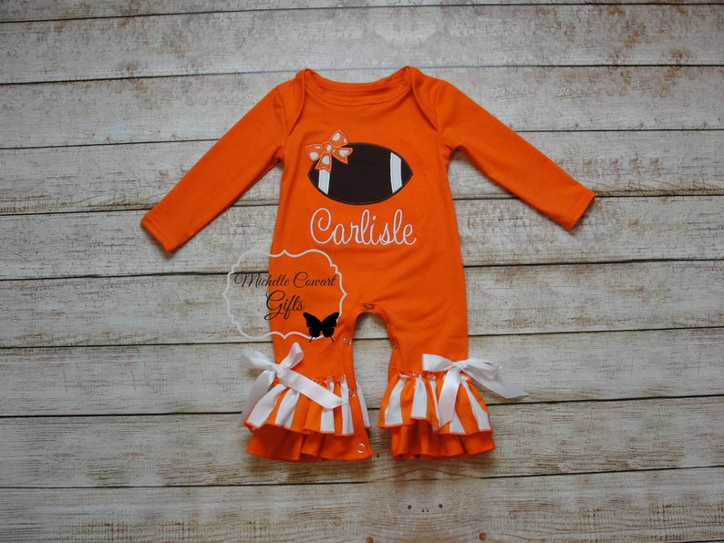 May include: Orange and white baby onesie with a football and bow design. The onesie has a ruffled bottom and says "Carlisle" on the front.