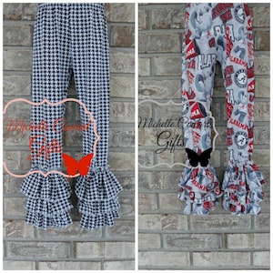 May include: Two pairs of children's pants. One pair features a black and white houndstooth pattern, and the other has a gray background with red and white Alabama-themed graphics. Both have ruffled hems.