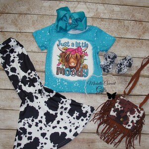 Pode incluir: Um conjunto infantil com uma camiseta turquesa com um desenho de vaca das Terras Altas e o texto "Just a little moody", cal&ccedil;as, sapatos, uma bolsa e um grande la&ccedil;o turquesa com estampas de vaca.