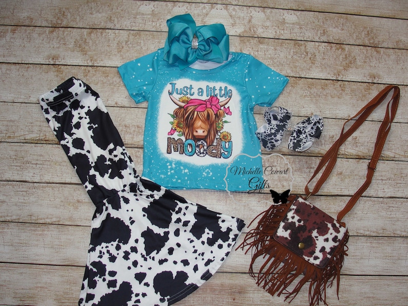 Pode incluir: Um conjunto infantil com uma camiseta turquesa com um desenho de vaca das Terras Altas e o texto "Just a little moody", cal&ccedil;as, sapatos, uma bolsa e um grande la&ccedil;o turquesa com estampas de vaca.