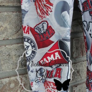 May include: White fabric with a repeating pattern of red and white collegiate logos, including the word "BAMA", a cartoon elephant, and pom-poms. The fabric is likely for clothing or accessories.