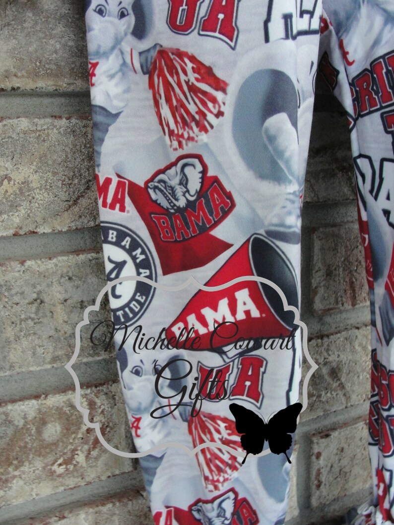 May include: White fabric with a repeating pattern of red and white collegiate logos, including the word "BAMA", a cartoon elephant, and pom-poms. The fabric is likely for clothing or accessories.