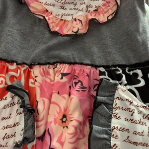 May include: A gray top with a floral skirt. The top has a ruffled collar with text. The skirt features a mix of floral patterns in pink, orange, and black, with ruffled accents.