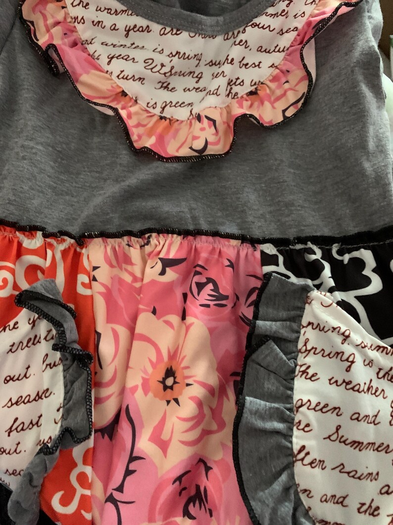 May include: A gray top with a floral skirt. The top has a ruffled collar with text. The skirt features a mix of floral patterns in pink, orange, and black, with ruffled accents.