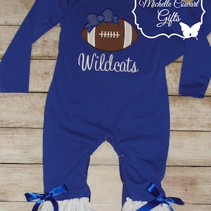 May include: A royal blue long-sleeved baby onesie with a white ruffle trim. The onesie features an embroidered football with a blue bow and the text "Wildcats".