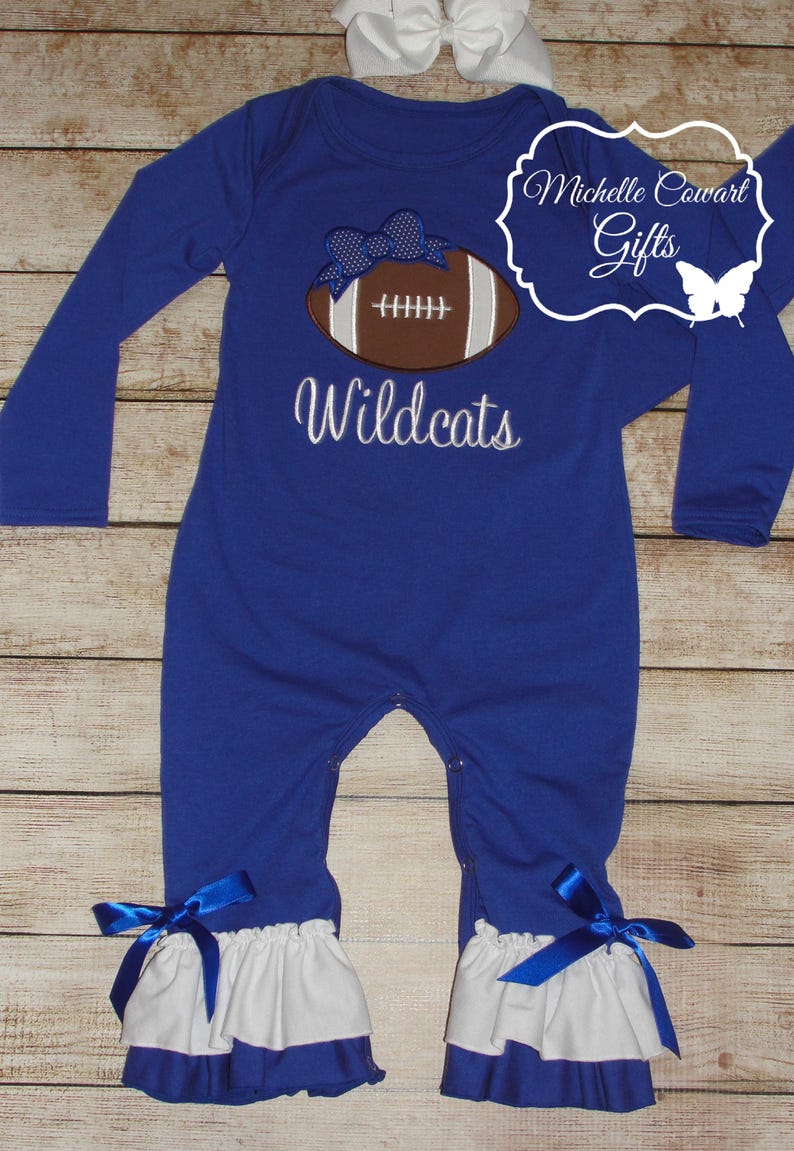 May include: A royal blue long-sleeved baby onesie with a white ruffle trim. The onesie features an embroidered football with a blue bow and the text "Wildcats".