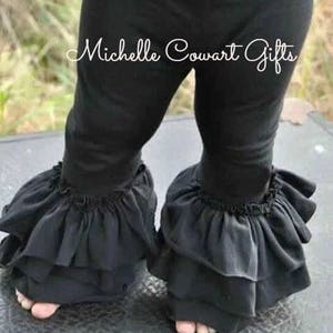 May include: Black leggings with ruffled trim at the ankles. The leggings are worn by a child. The text "Michelle Cowart Gifts" is visible on the leggings. The leggings are made of a soft material.