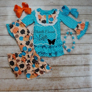 Puede incluir: Conjunto turquesa de lunares para ni&ntilde;a, con una blusa de manga larga con detalles de calabazas y volantes, combinada con pantalones a juego con estampado de calabazas. Incluye lazos naranjas y azules, y un collar de perlas. El texto "Michelle Cowart Gifts" es visible.