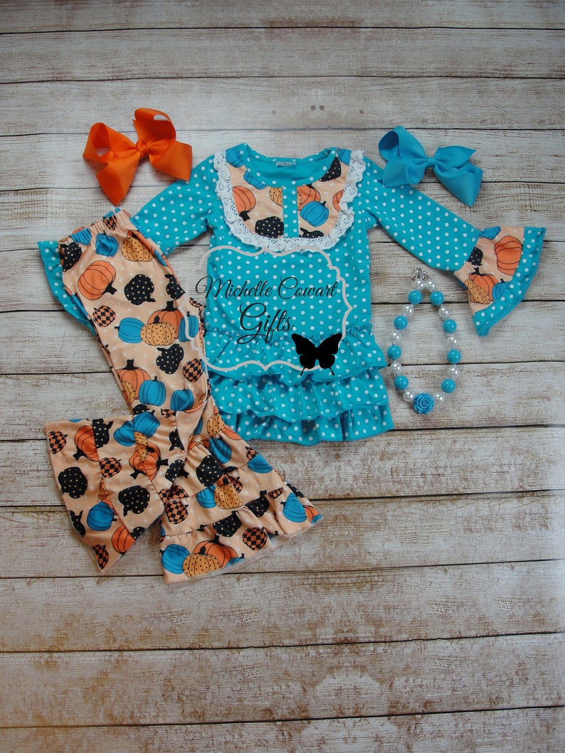 Puede incluir: Conjunto turquesa de lunares para ni&ntilde;a, con una blusa de manga larga con detalles de calabazas y volantes, combinada con pantalones a juego con estampado de calabazas. Incluye lazos naranjas y azules, y un collar de perlas. El texto "Michelle Cowart Gifts" es visible.