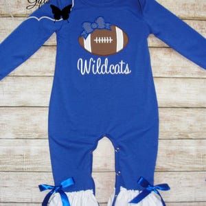 May include: A royal blue baby onesie with long sleeves and a white ruffle trim. It features an embroidered brown football with a blue bow and the text "Wildcats".