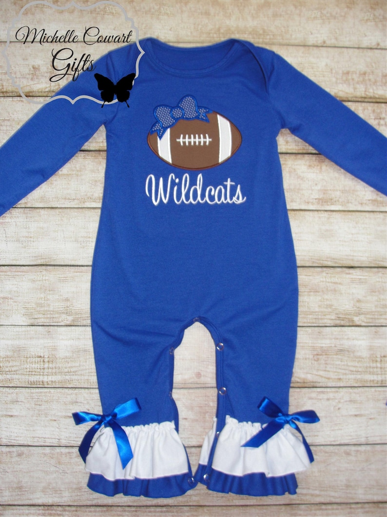 May include: A royal blue baby onesie with long sleeves and a white ruffle trim. It features an embroidered brown football with a blue bow and the text "Wildcats".
