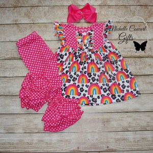 Puede incluir: Un conjunto de tres piezas para ni&ntilde;a beb&eacute; que incluye leggings rosas con lunares y volantes en la parte inferior, una blusa rosa con lunares con una falda de estampado de arco&iacute;ris y leopardo, y una diadema rosa con lazo.  Michelle Cowart Gifts.