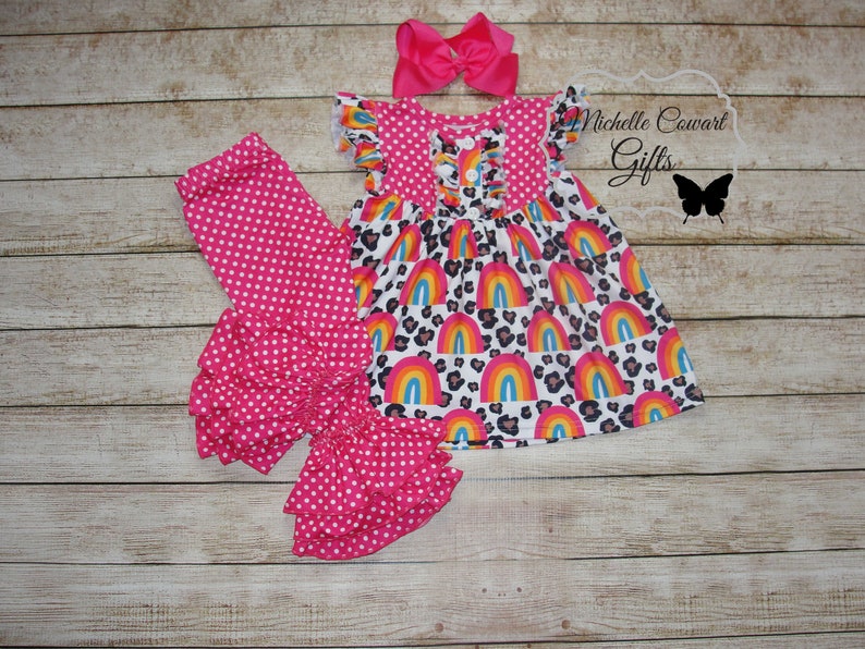 Puede incluir: Un conjunto de tres piezas para ni&ntilde;a beb&eacute; que incluye leggings rosas con lunares y volantes en la parte inferior, una blusa rosa con lunares con una falda de estampado de arco&iacute;ris y leopardo, y una diadema rosa con lazo.  Michelle Cowart Gifts.