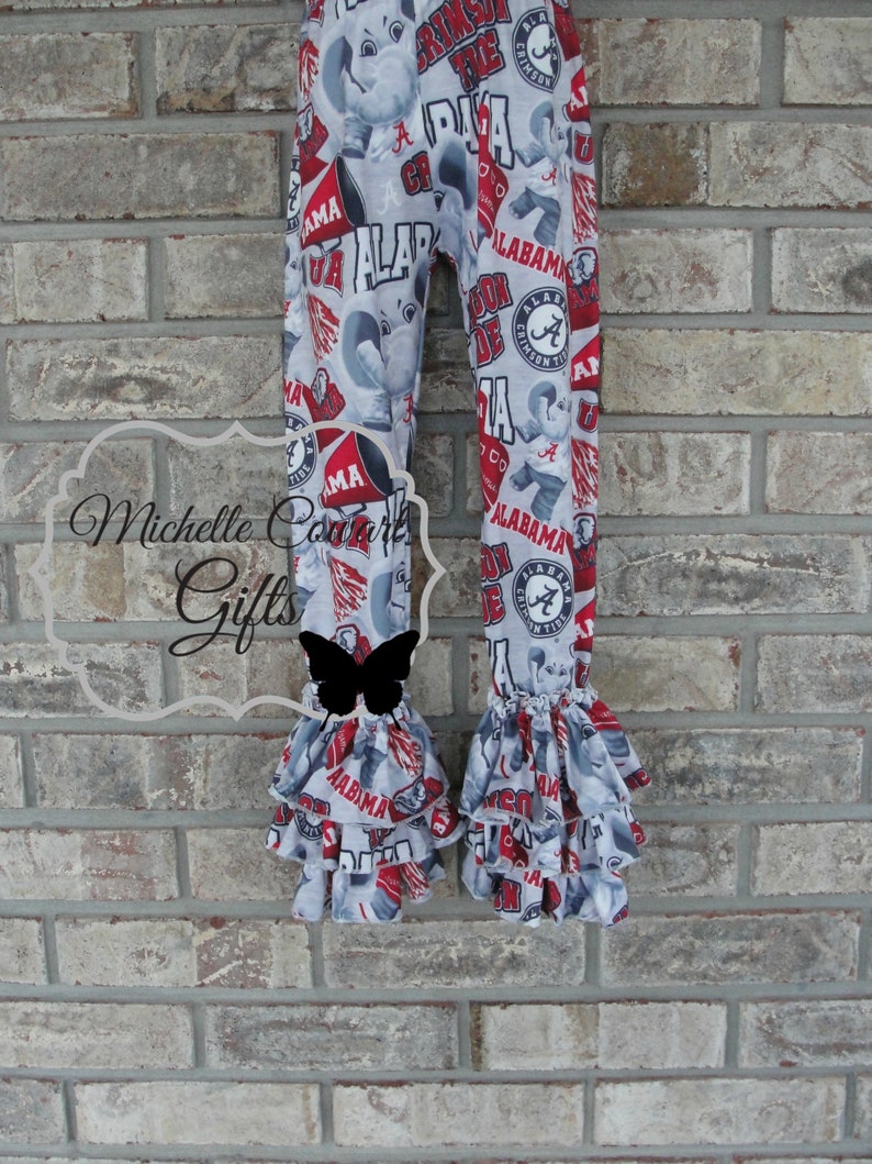 May include: Gray and red patterned leggings with ruffles at the ankles. The leggings feature the word "ALABAMA" and the Crimson Tide logo. The fabric has a repeating pattern of elephants and team-related graphics.