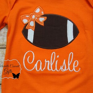 May include: Orange long-sleeved shirt with a brown and white football design and a white bow with orange and white polka dots. The shirt is personalized with the name "Carlisle" in white cursive.