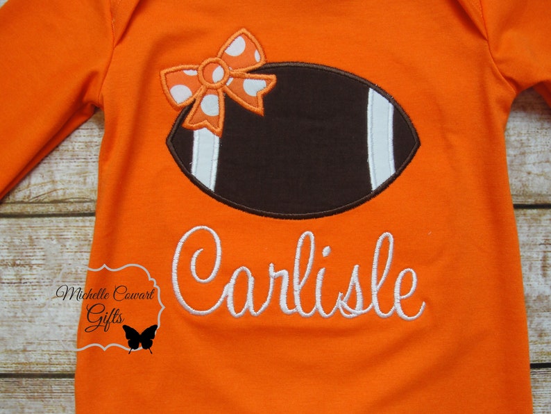 May include: Orange long-sleeved shirt with a brown and white football design and a white bow with orange and white polka dots. The shirt is personalized with the name "Carlisle" in white cursive.