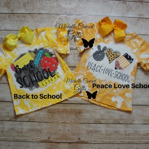 Op de afbeelding: Twee gele tie-dye shirts met verschillende ontwerpen voor de terugkeer naar school. E&eacute;n shirt zegt "Back to School" met een potlood, een liniaal en een appel. Het andere shirt zegt "Peace Love School" met een vredesteken, een hart en schoolbenodigdheden.