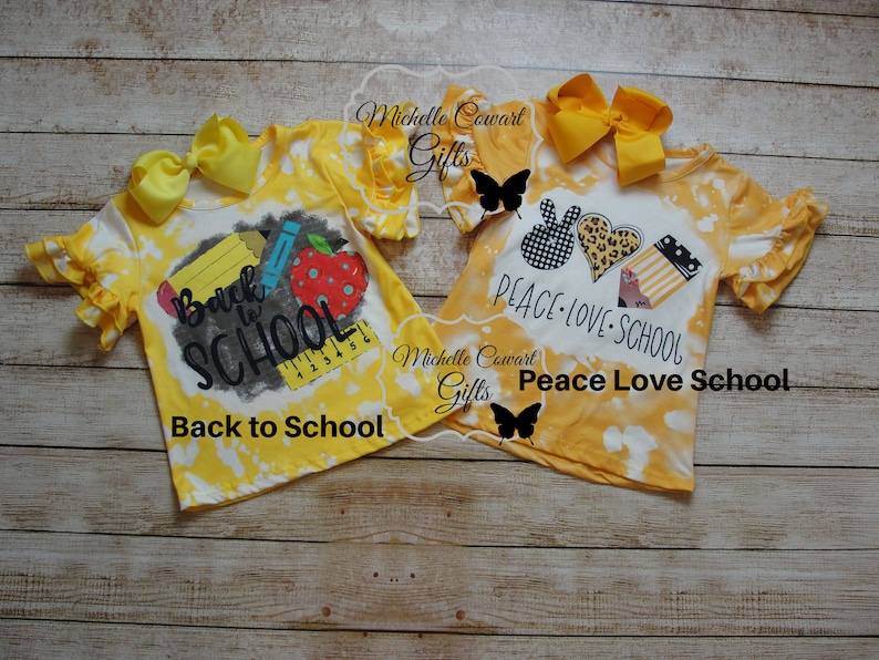 Op de afbeelding: Twee gele tie-dye shirts met verschillende ontwerpen voor de terugkeer naar school. E&eacute;n shirt zegt "Back to School" met een potlood, een liniaal en een appel. Het andere shirt zegt "Peace Love School" met een vredesteken, een hart en schoolbenodigdheden.