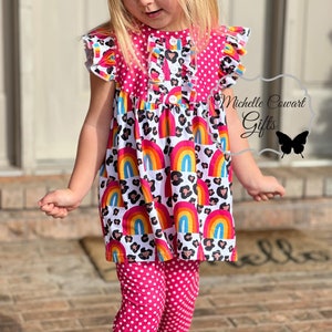 Puede incluir: Una ni&ntilde;a peque&ntilde;a lleva un vestido de lunares rosa y blanco con un dise&ntilde;o de arco&iacute;ris y estampado de leopardo. El vestido tiene un borde de volantes y un cierre de bot&oacute;n. Tambi&eacute;n lleva pantalones de lunares rosa con un borde de volantes en la parte inferior.